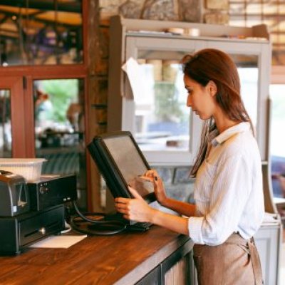 A hospitality worker stands at the counter of a cafe, typing into a computer.